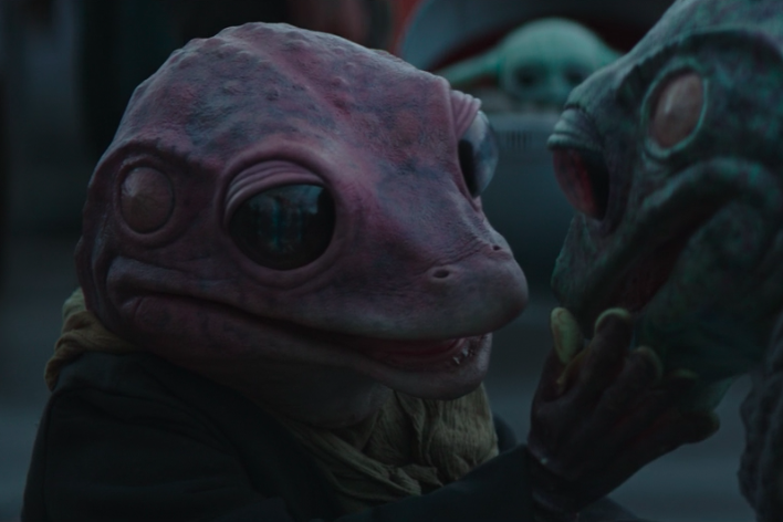 A reptilian creature with large, black eyes and bumpy pink puts her hand to the cheek of a similar creature who has green skin.