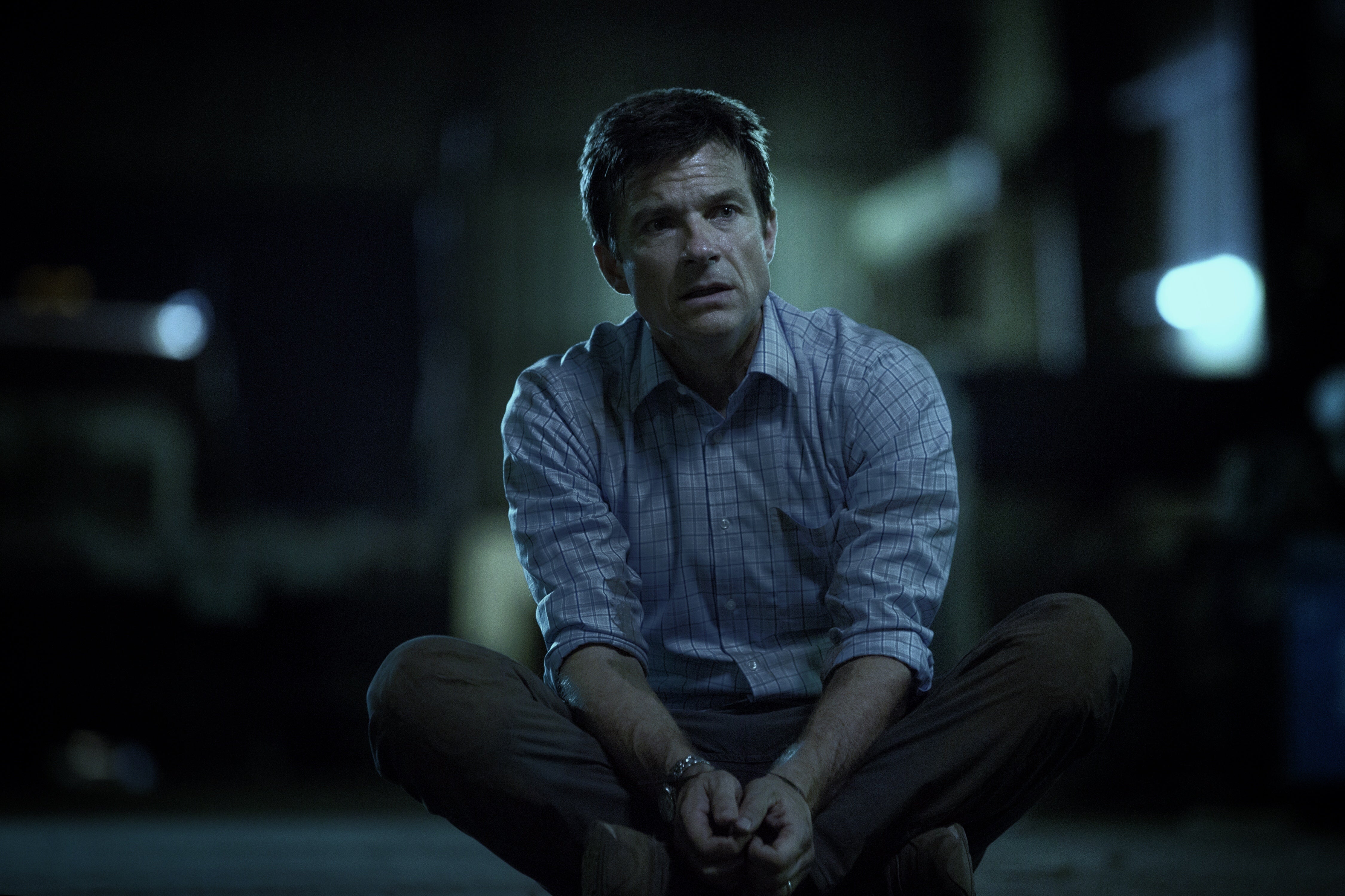 Jason Bateman sits cross-legged with his hands in front of him.