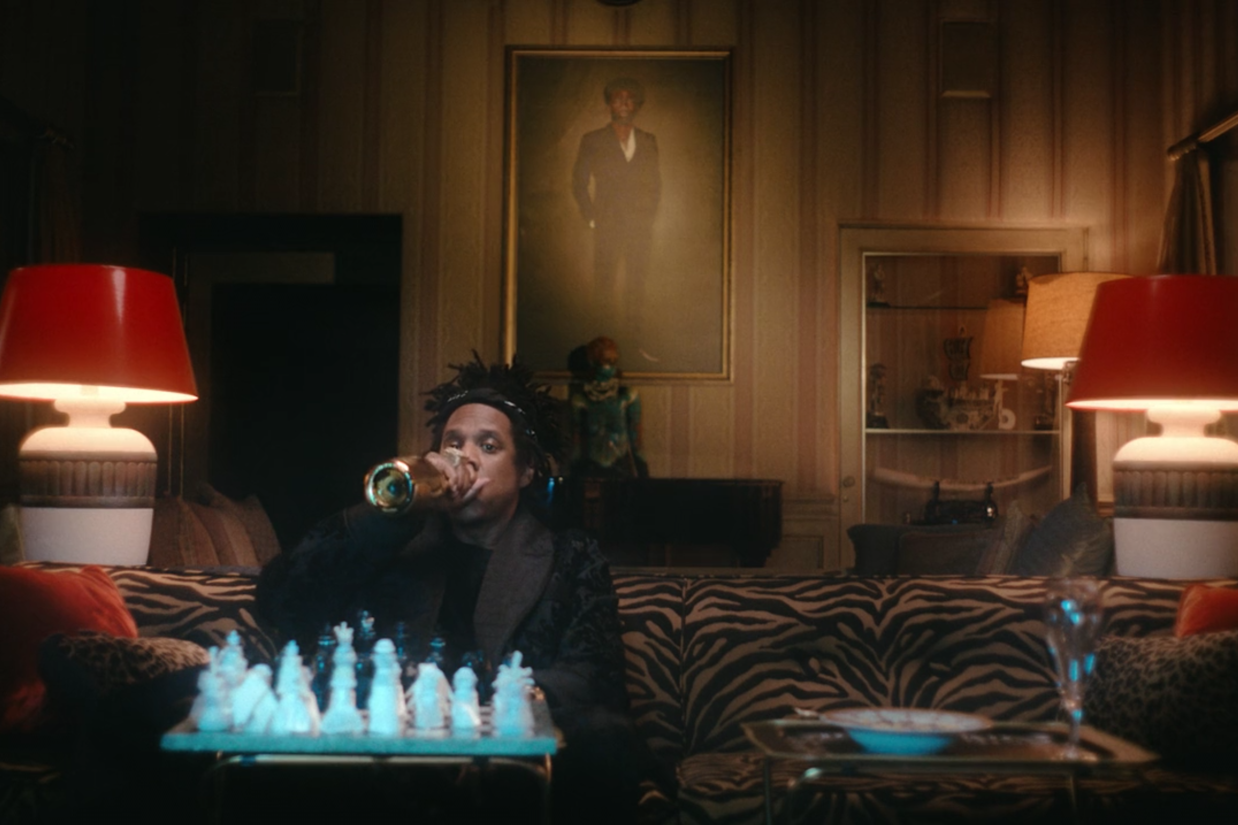 Jay-Z drinks from a gold bottle while sitting in front of a chessboard.