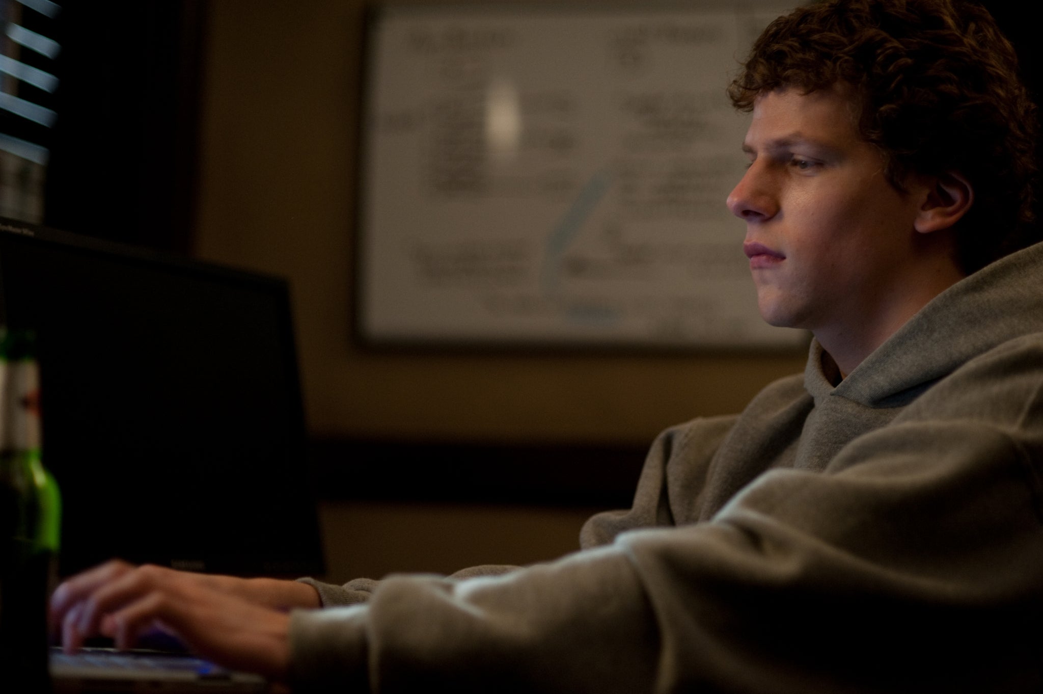 Jesse Eisenberg portrays a young Mark Zuckerberg in his college dorm room in a plain grey sweatshirt while he types on his laptop. 