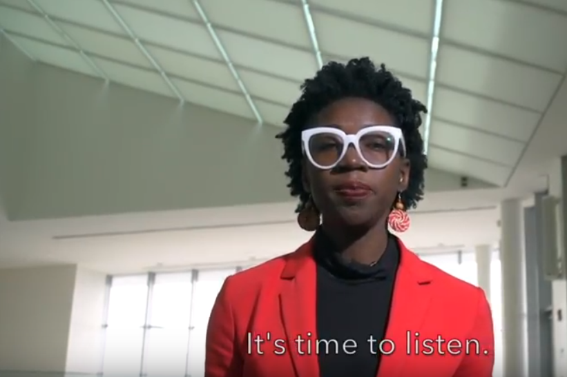 Joy Buolamwini wears white glasses, a black shirt, and a red jacket while standing in a hallway.
