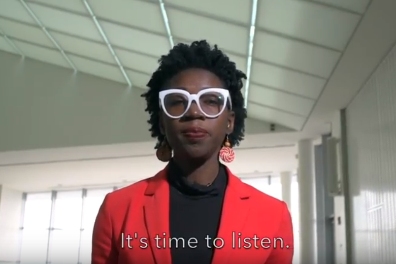 Joy Buolamwini wears white glasses, a black shirt, and a red jacket while standing in a hallway.