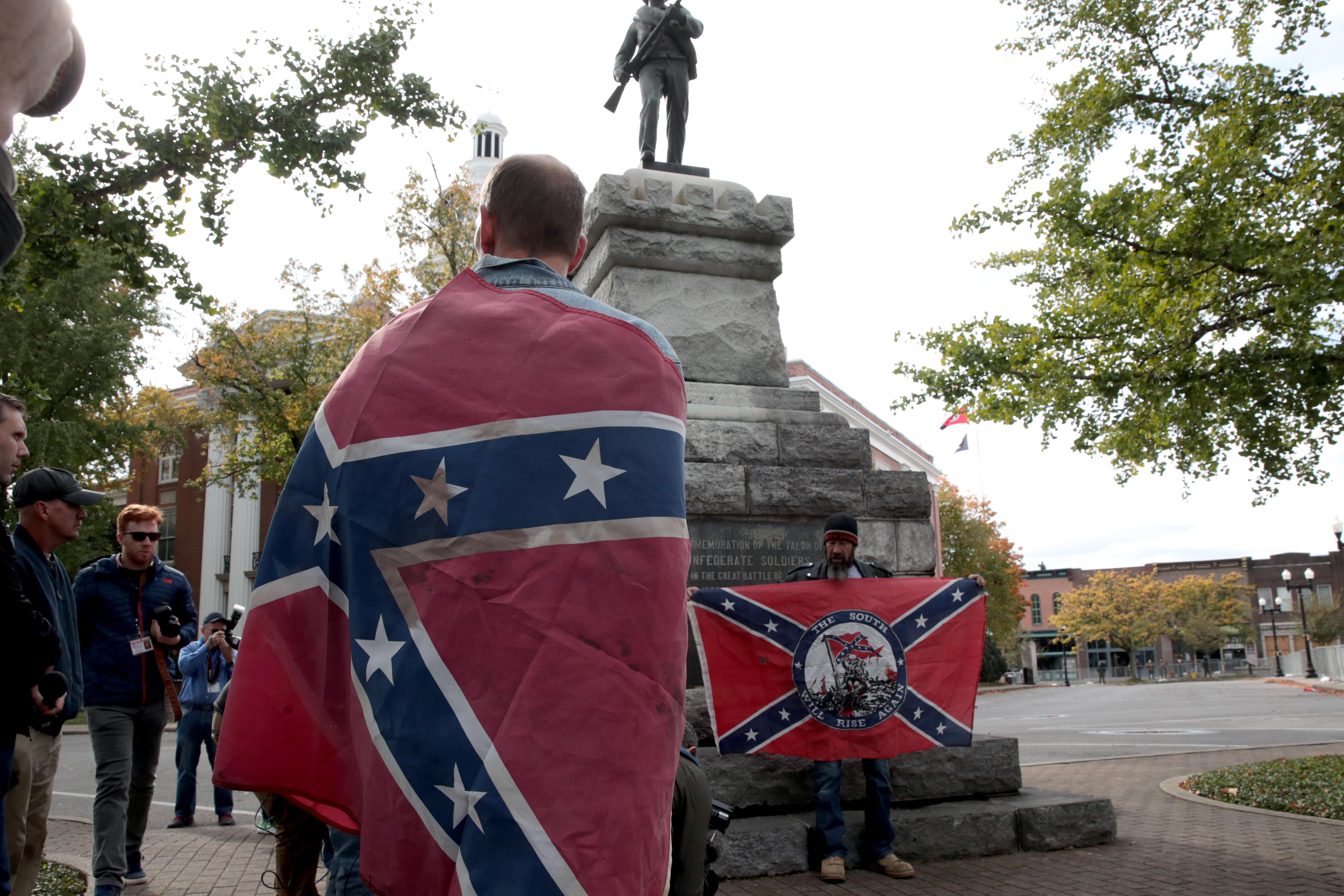 A man wrapped in a Confederate flag stands in front of a Confederate statue at a rally.