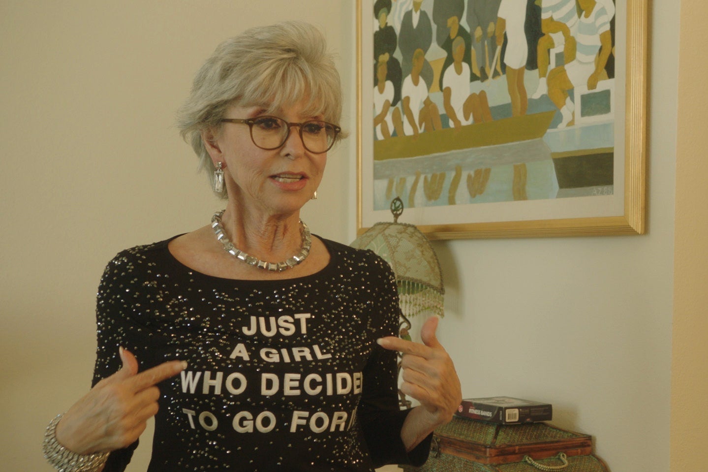 Rita Moreno, with gray hair and glasses, points her black, bedazzled shirt. Text on the shirt reads: "Just A Girl Who Decided to Go For It"