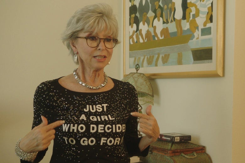 Rita Moreno, with gray hair and glasses, points her black, bedazzled shirt. Text on the shirt reads: "Just A Girl Who Decided to Go For It"