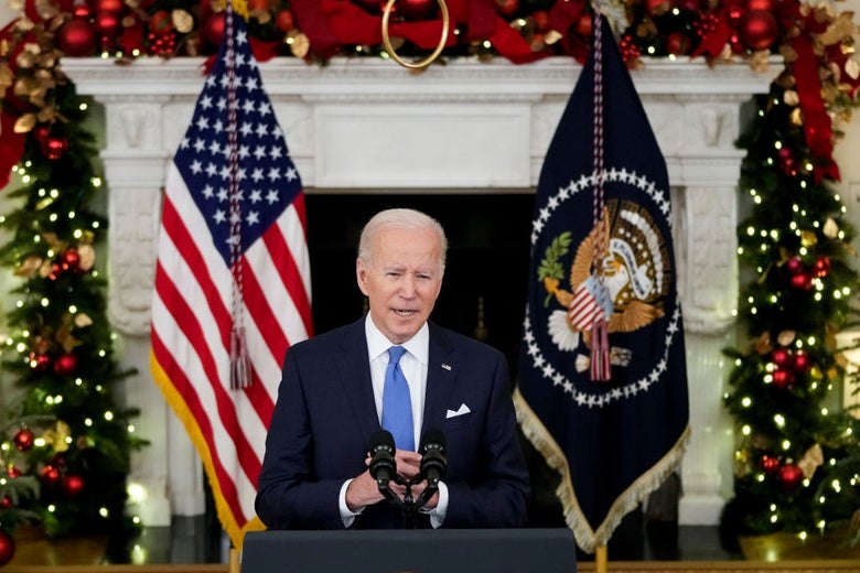 Biden speaks from a lectern in the White House in front of a backdrop that includes two Christmas trees and a mantle decorated with ribbons and ornaments.