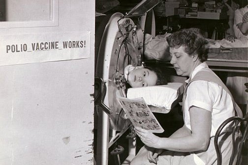A sign reads, "Polio vaccine works," beside a girl in an iron lung with a woman reading to her.