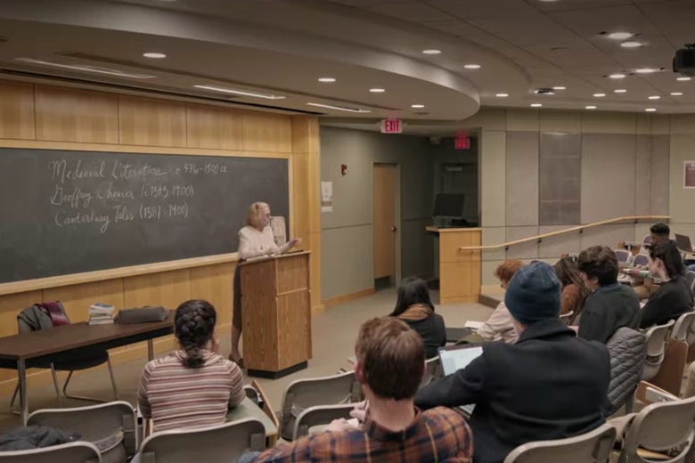 Joan teaching from a podium in a well-lit college classroom with rows of gray plastic chairs in it.