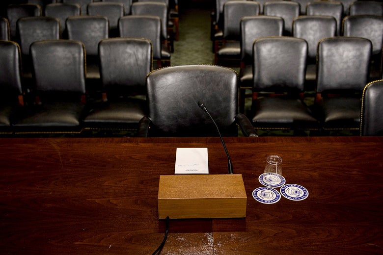 Empty chair behind the desk in the hearing room, with rows of empty chairs behind it.
