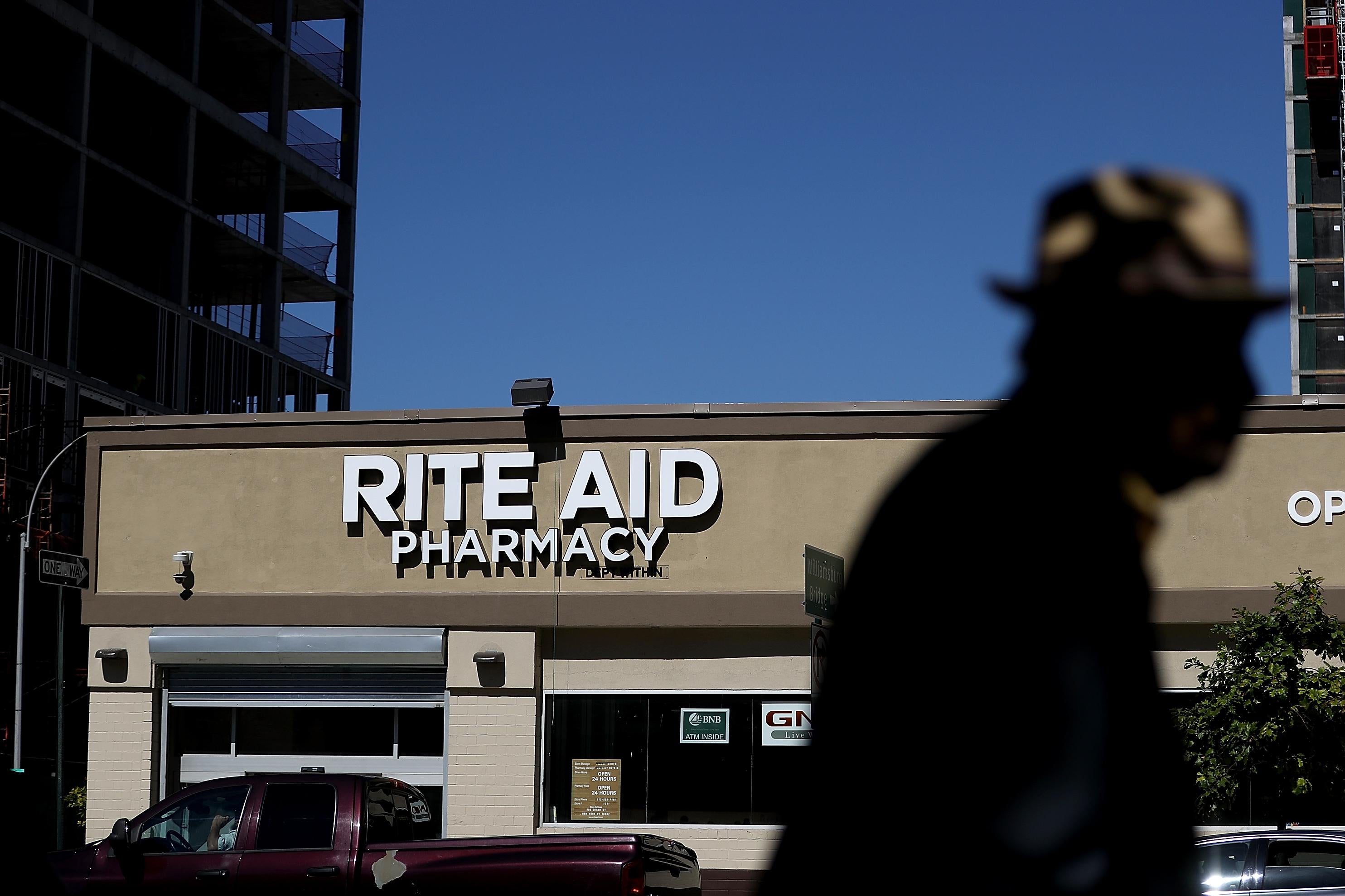 A pedestrian, silhouetted, walks by a dark red pick up truck and a beige storefront with a white sign that reads, "Rite Aid Pharmacy."