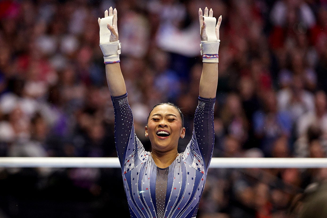 Lee beams with her eyes closed and arms outstretched into the air with the uneven bars and the crowd behind her