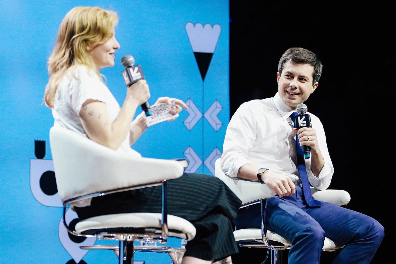 Buttigieg, wearing a white shirt and blue tie, laughs as Cox speaks on stage against a blue background.