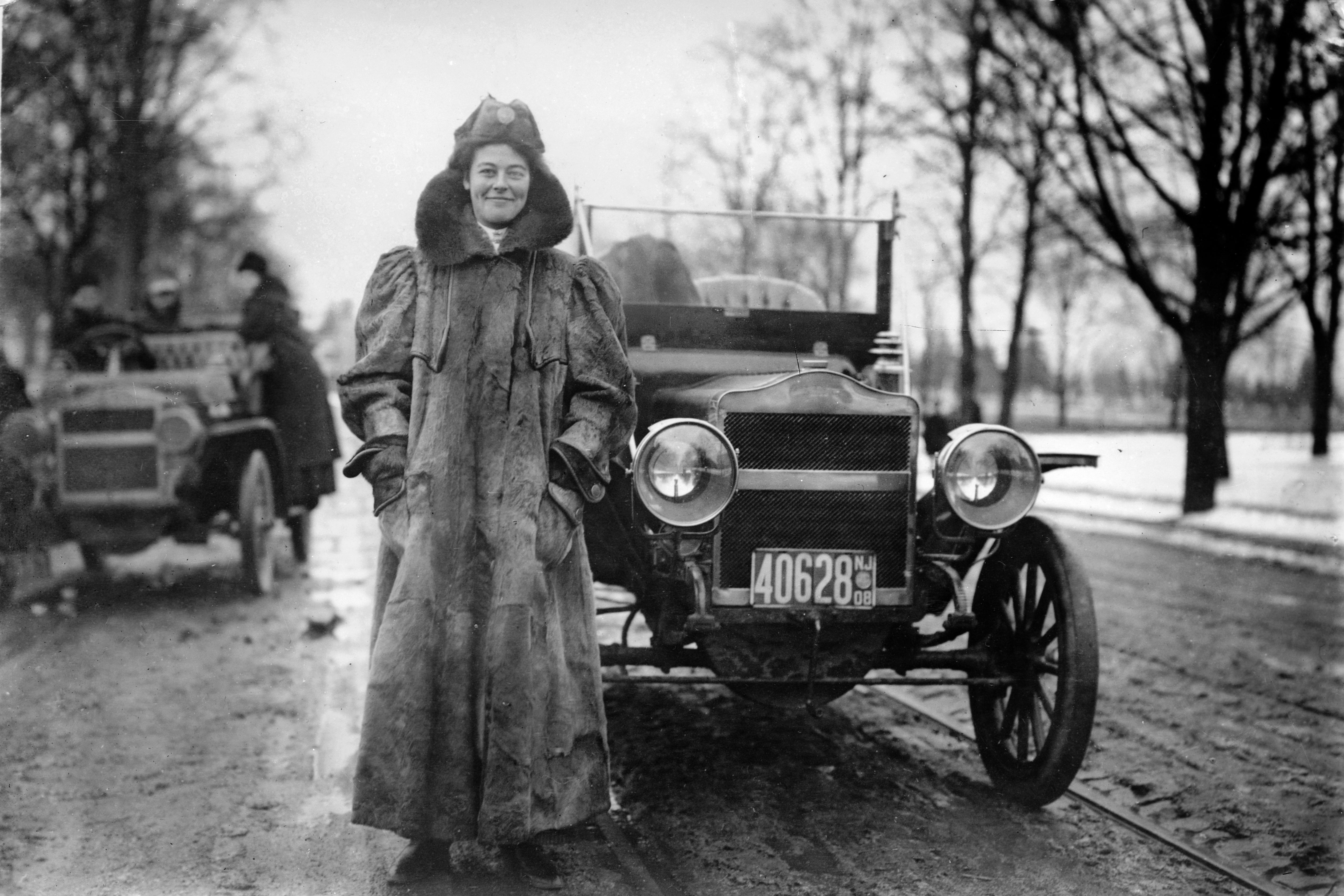 A woman in a full-length coat stands smiling in front of an old-timey car.