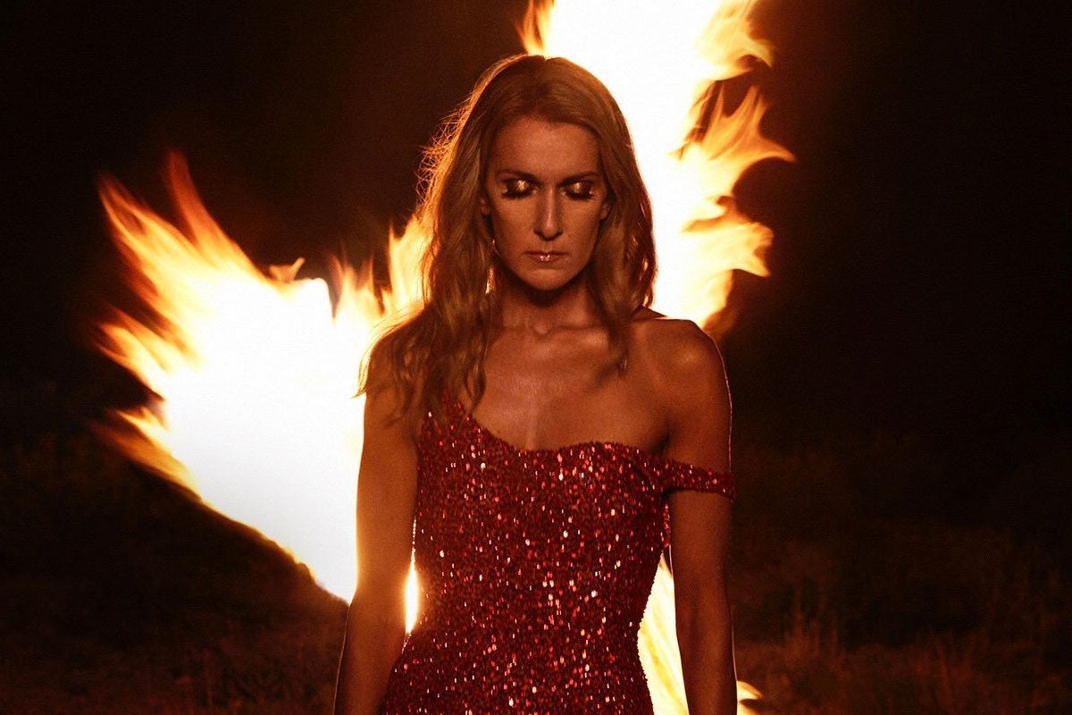 Celine Dion looks downward as a fire rages behind her.