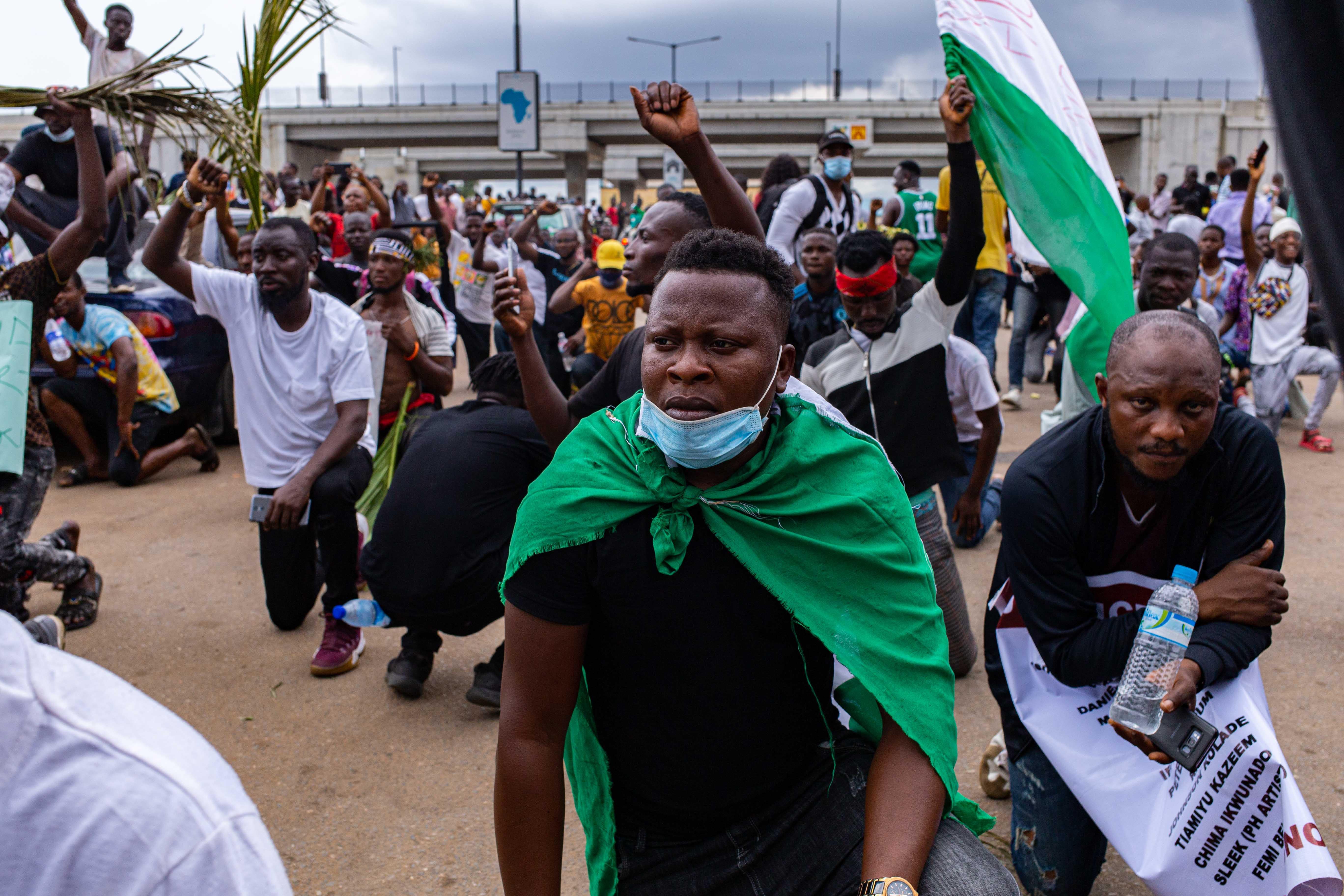 A man wearing a Nigerian flag and a face mask takes a knee while others raise their fists as a sign of protest. 