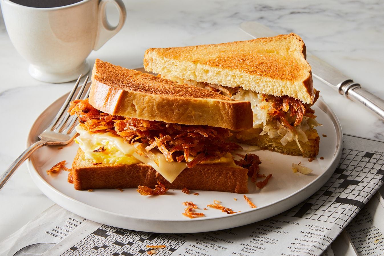Latke, egg, and cheese sandwich sliced into triangles on a plate with a newspaper crossword folded under it and a coffee cup on the table beside it.