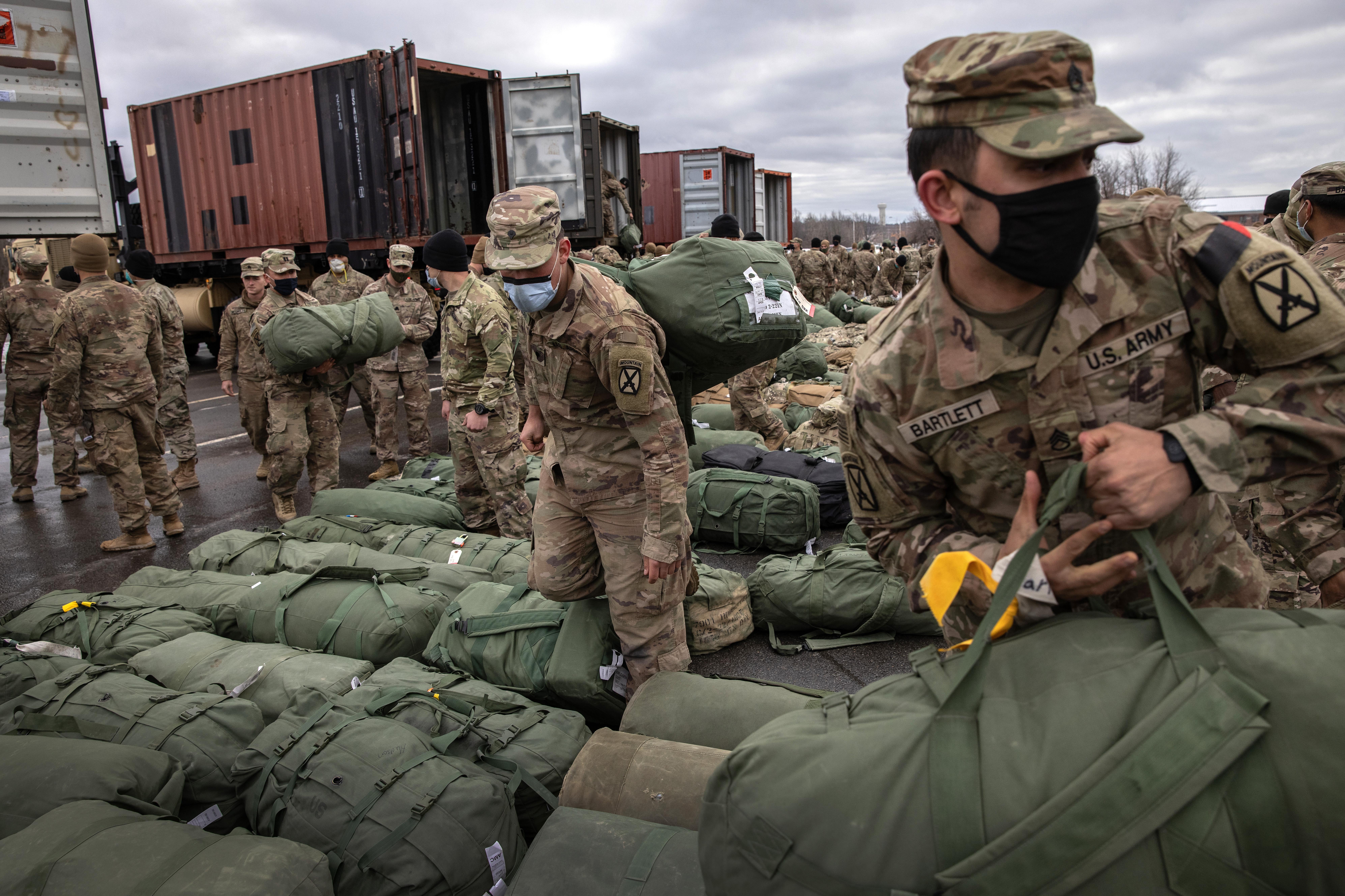 Soldiers in fatigues pick up their duffel bags, which are arranged in neat rows on the ground. There are shipping containers in the background.