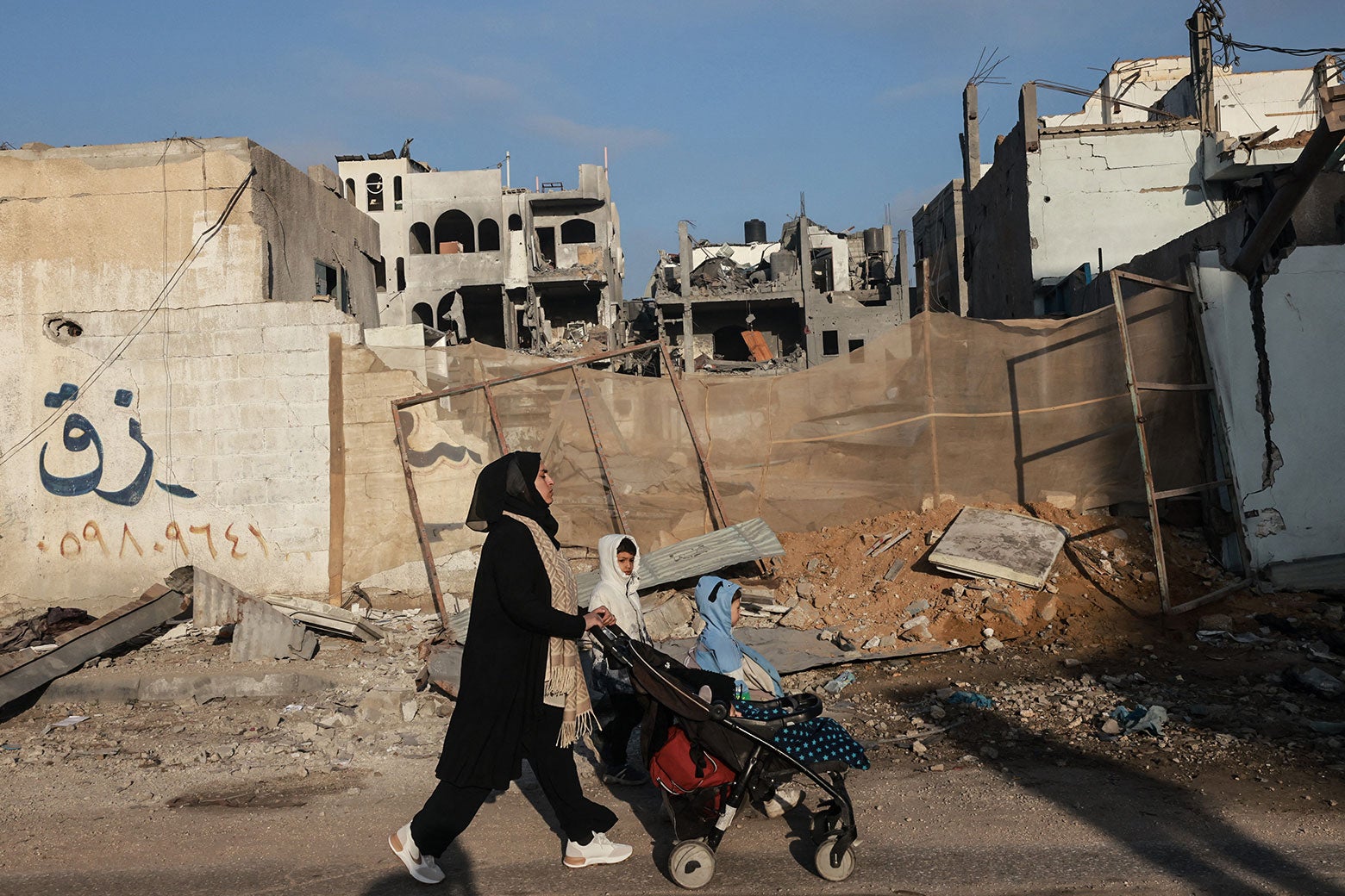 A woman walking with two small children pushes a stroller past ruined buildings.