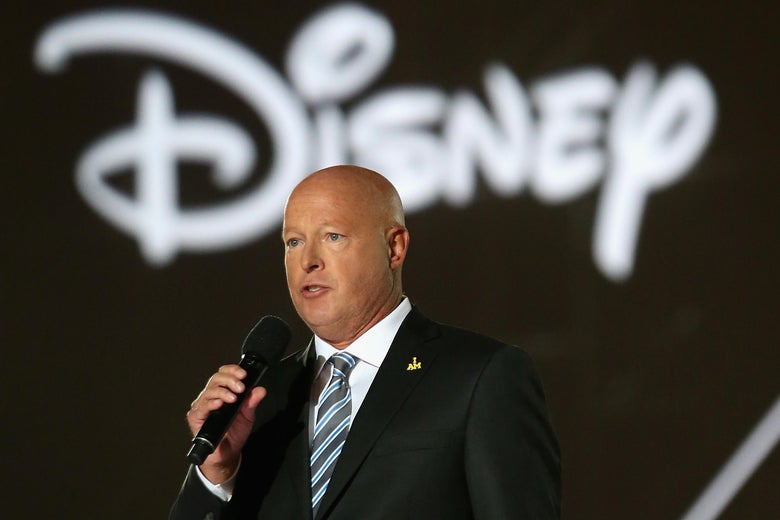 Bob Chapek, a man in a suit, stands before a sign reading "Disney," holding a microphone.