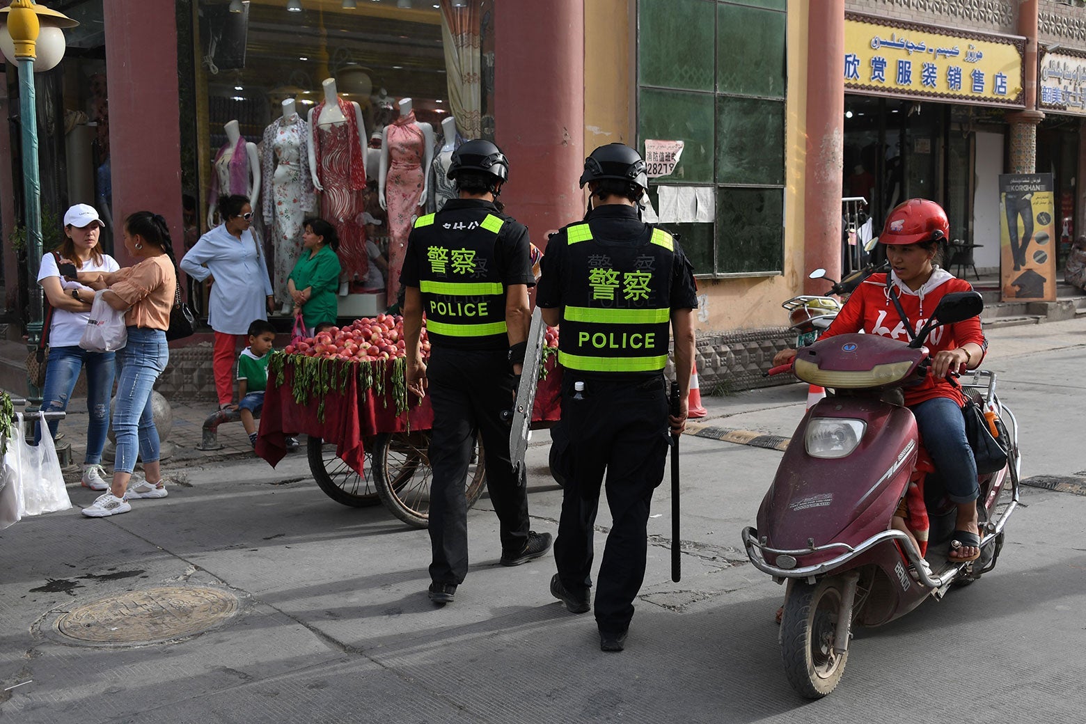 Police officers walk along a street near a fruit cart as a moped passes.