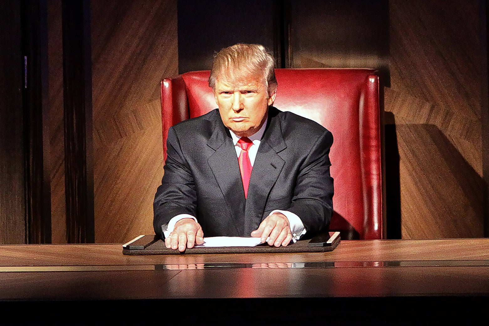 Trump sits in a red boardroom chair looking very serious and squinting as if thinking very hard.