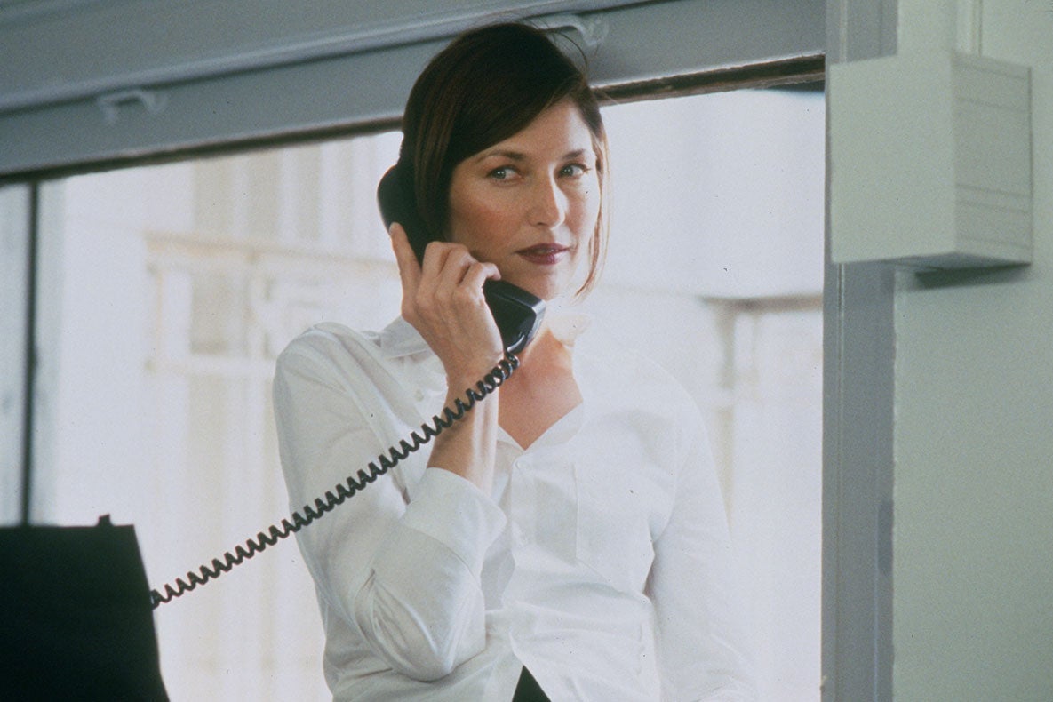 A woman in a white button-down shirt holds a landline phone to her ear while standing in a drab office
