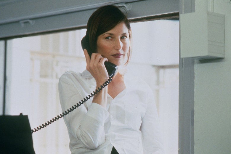 A woman in a white button-down shirt holds a landline phone to her ear while standing in a drab office