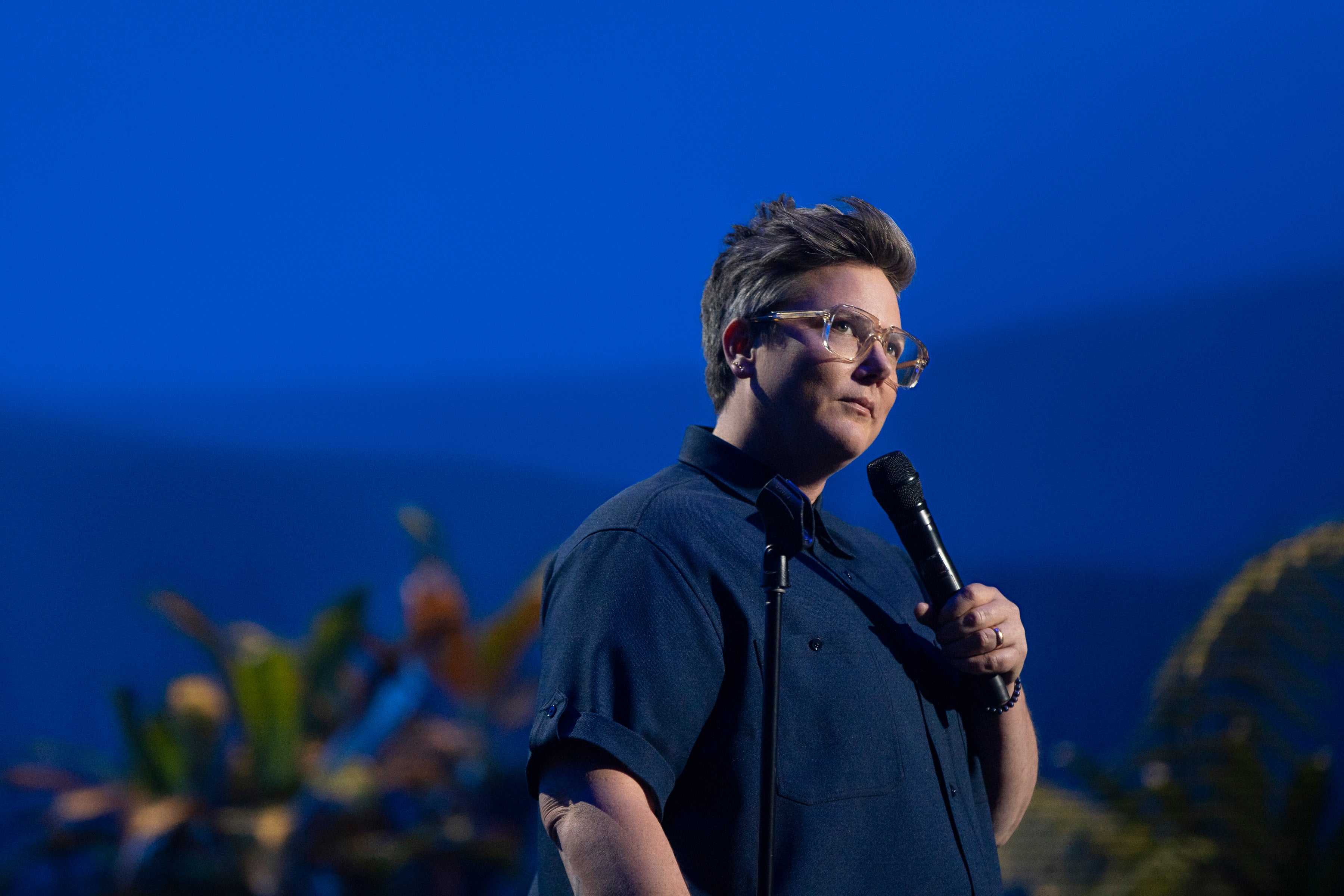 The comedian stands in front of a blue background and some rather tropical-looking plants, wearing a large navy-blue buttondown. They pause and gaze up at the folks in the higher seats.