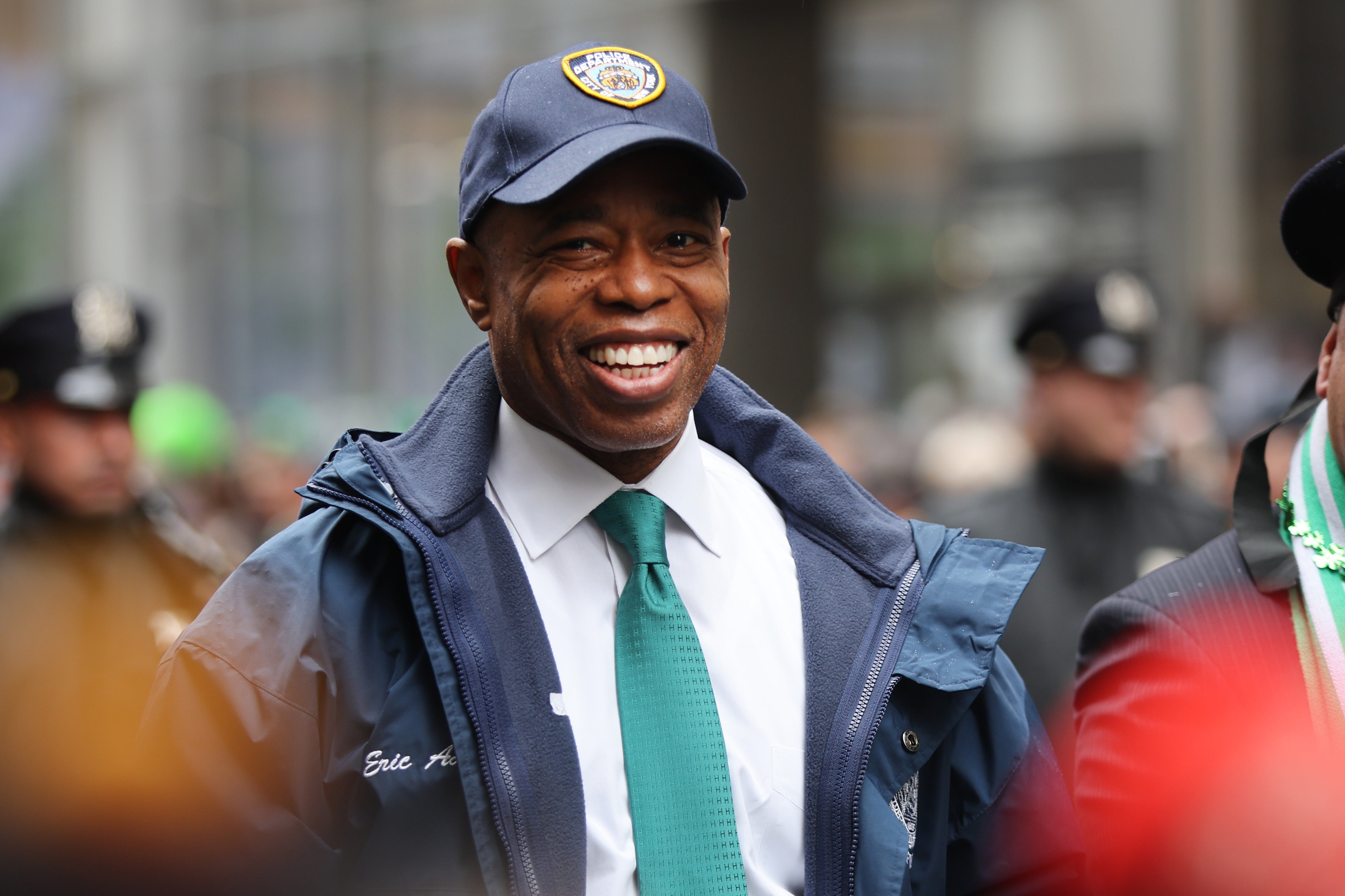 New York City mayor Eric Adams, wearing a green tie and an NYPD hat, smiles as he walks in a St. Patrick's Day parade.