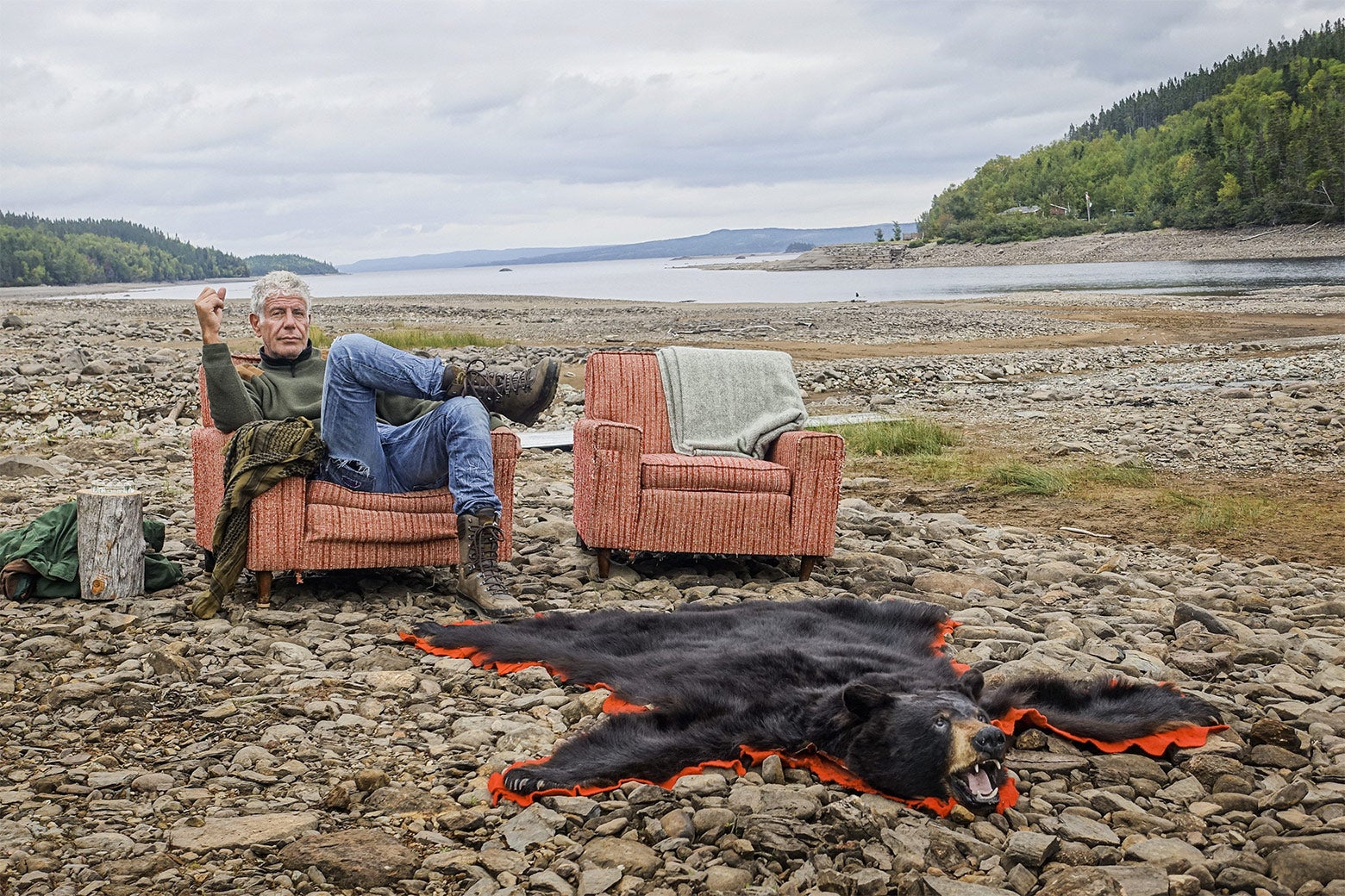 Anthony Bourdain sits in an upholstered chair on a rocky beach next to a bear rug.