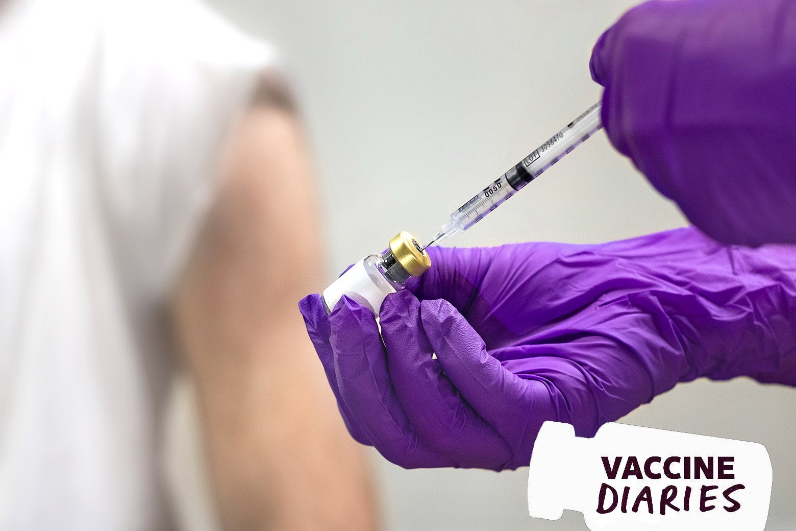 In the foreground, a purple-gloved hand drawing vaccine out of a vial. In the background, a person with their sleeve rolled up to expose their arm.