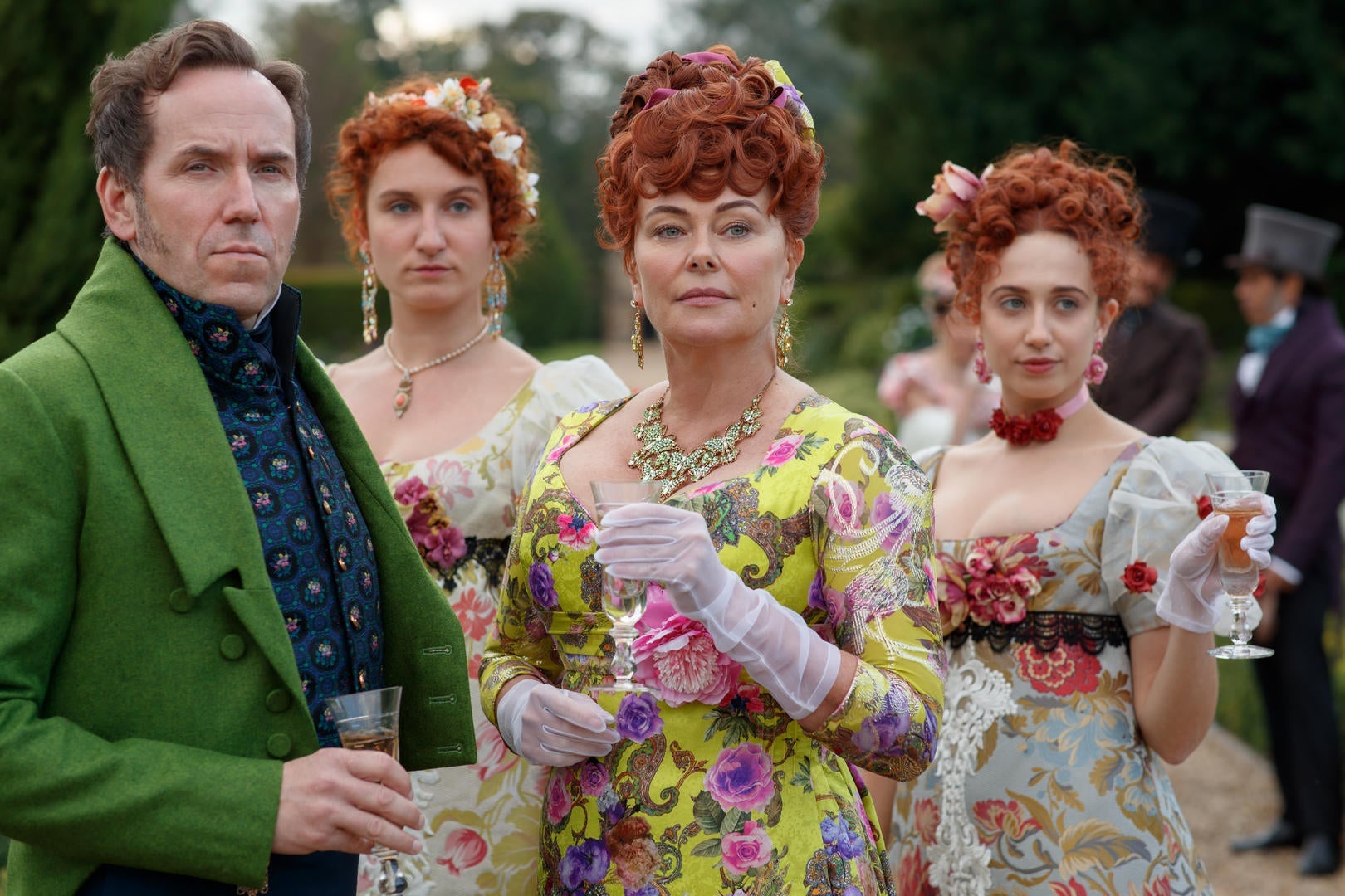 Ben Miller as Lord Featherington, wearing a bright green coat, stands with Polly Walker, who wears a yellow dress with a busy pattern of pink and purple flowers. Behind them, Harriet Cains as Philippa wears a low-cut floral dress with ruffled sleeves.