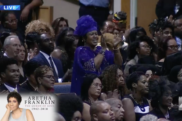 A woman in a purple ruffled hat and a purple jeweled top stands and claps in the audience of Aretha Franklin's funeral