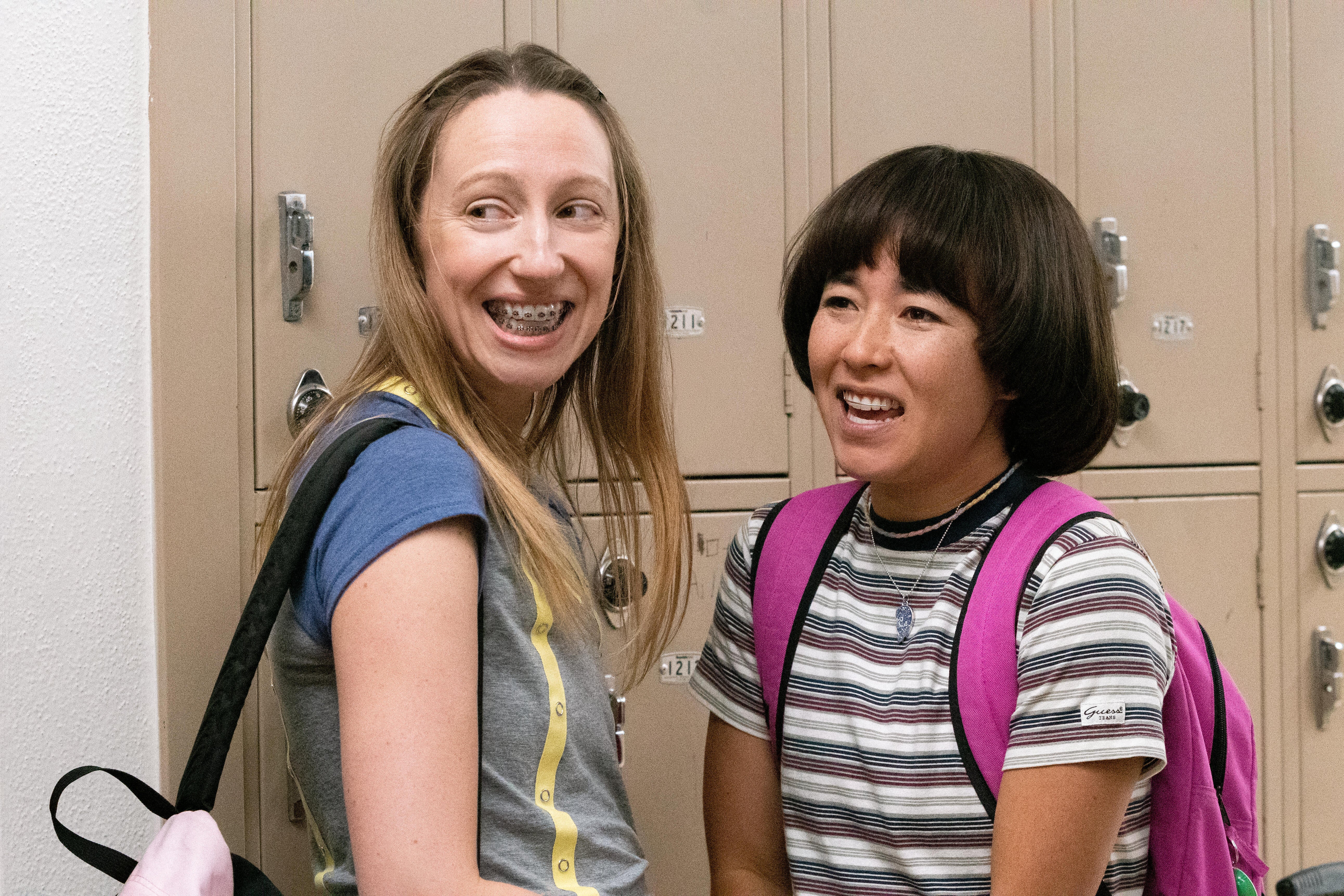 Two teenage girls, one with braces, smile awkwardly in a middle-school hallway.