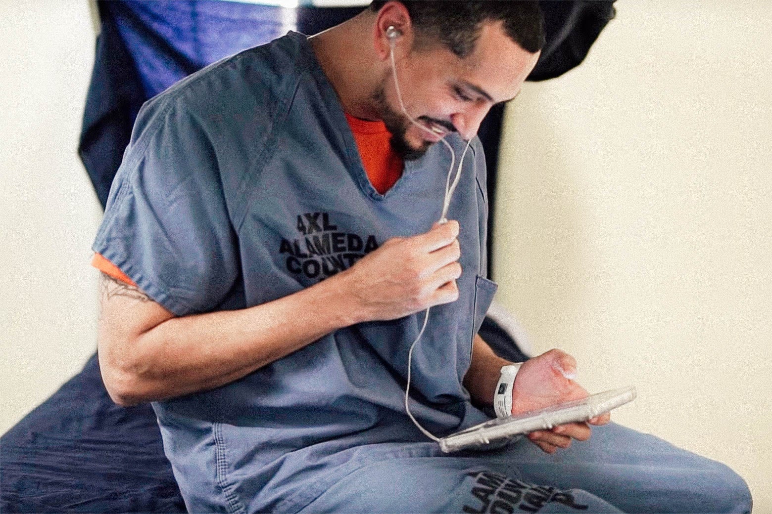 An inmate sits on a cot, smiling as he uses a GTL tablet with earbuds.