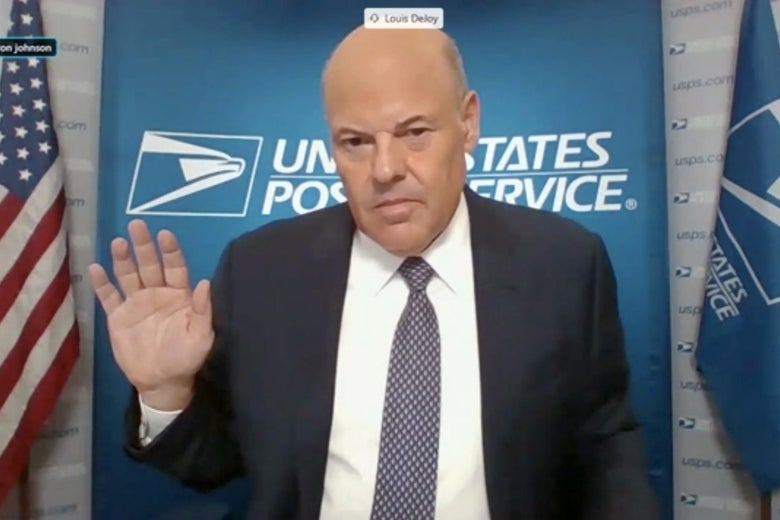Louis DeJoy raises his hand in front of a USPS backdrop and flag.