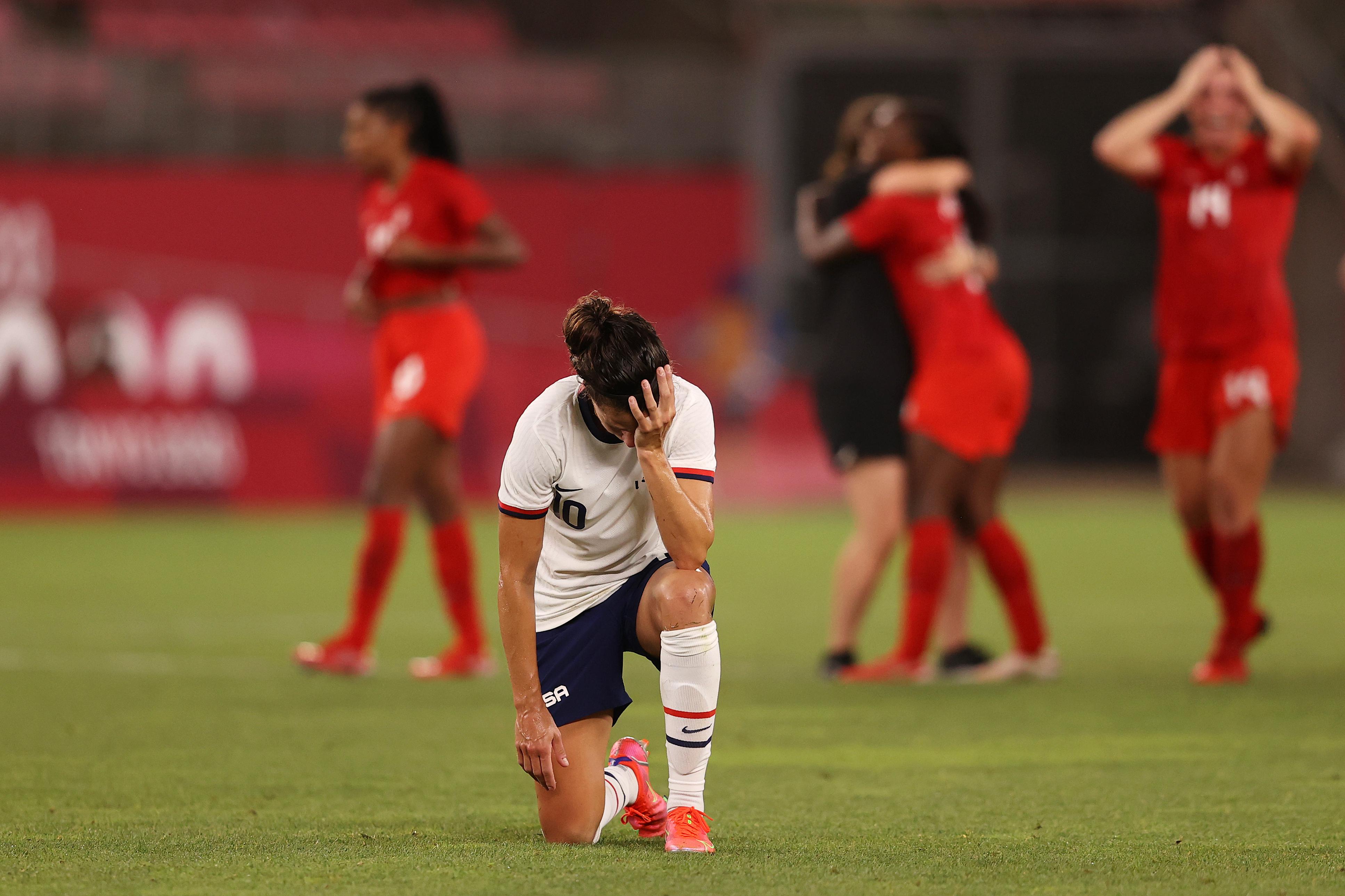 Lloyd on one knee with her head in her hand, looking dejected, as Canadian players celebrate behind her.
