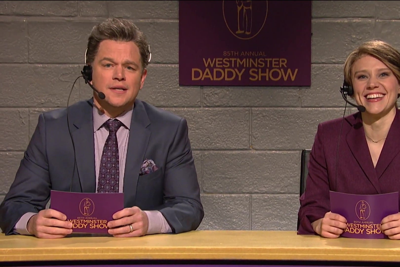 Matt Damon and Kate McKinnon sit at the announcer’s podium for the Westminster Daddy Show.