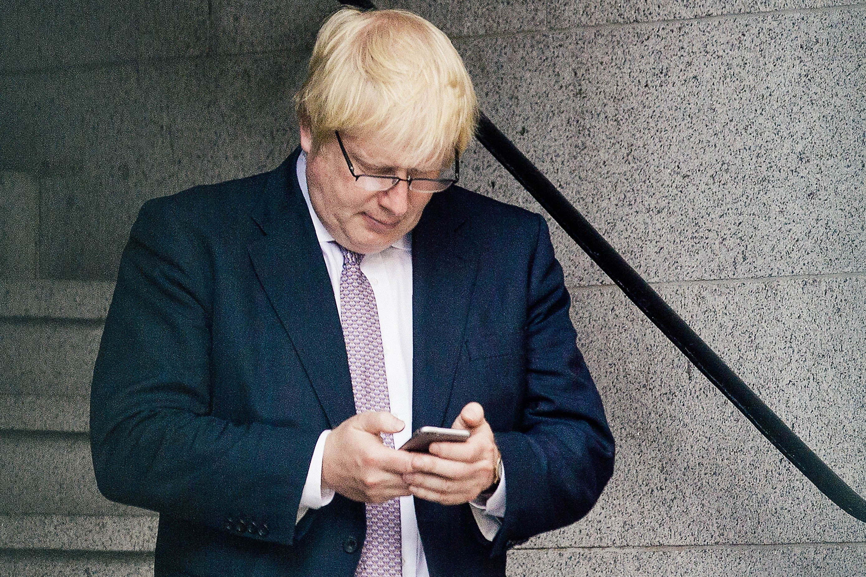 Boris Johnson looks down at a smartphone with his glasses on and a pensive expression.