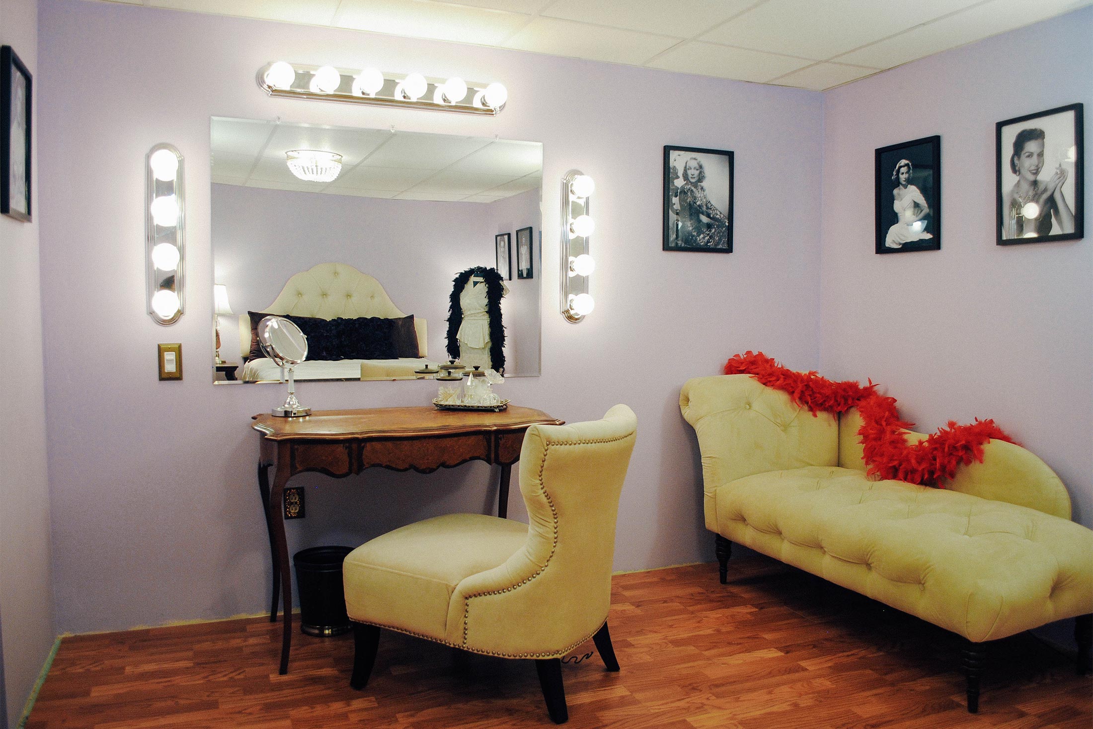 A lavender room with a vanity surrounded by globe lights, and a chaise draped in a red feather boa.