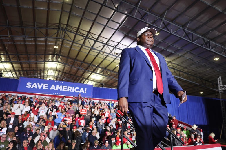 Gibbs walks out on stage in a white baseball cap and blue suit. The words "SAVE AMERICA!" appear behind him over a crowd of rallygoers.