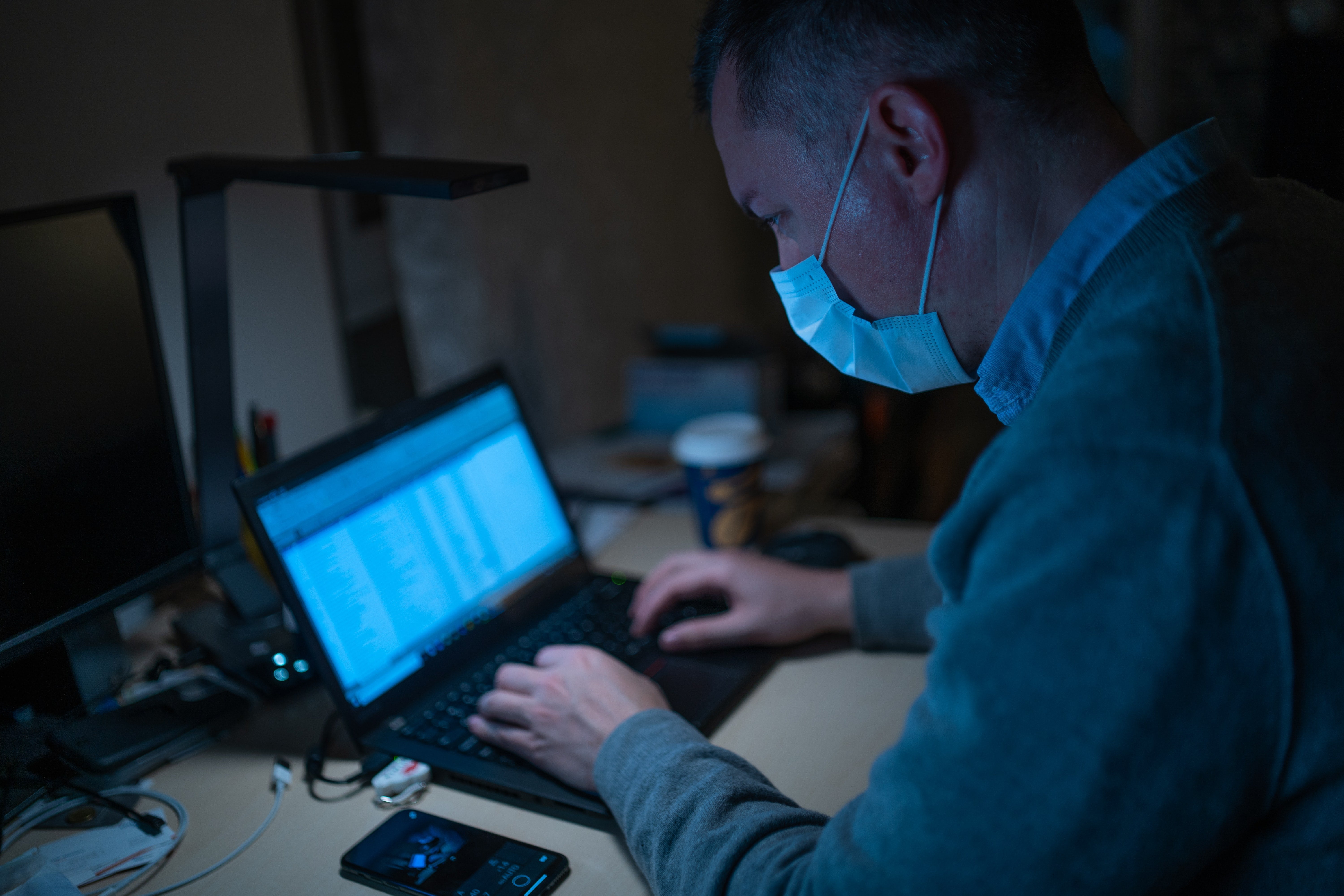 A man wearing a surgical mask types on a laptop at a desk.