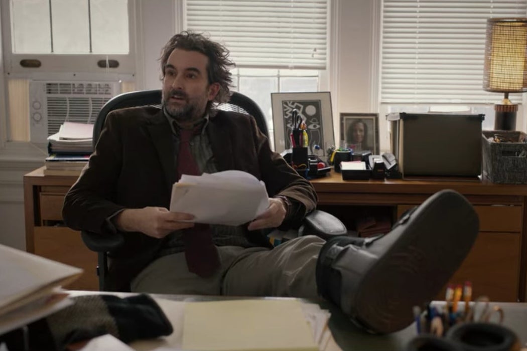 Bill sits on a chair in his office with one leg, injured, up on his desk.