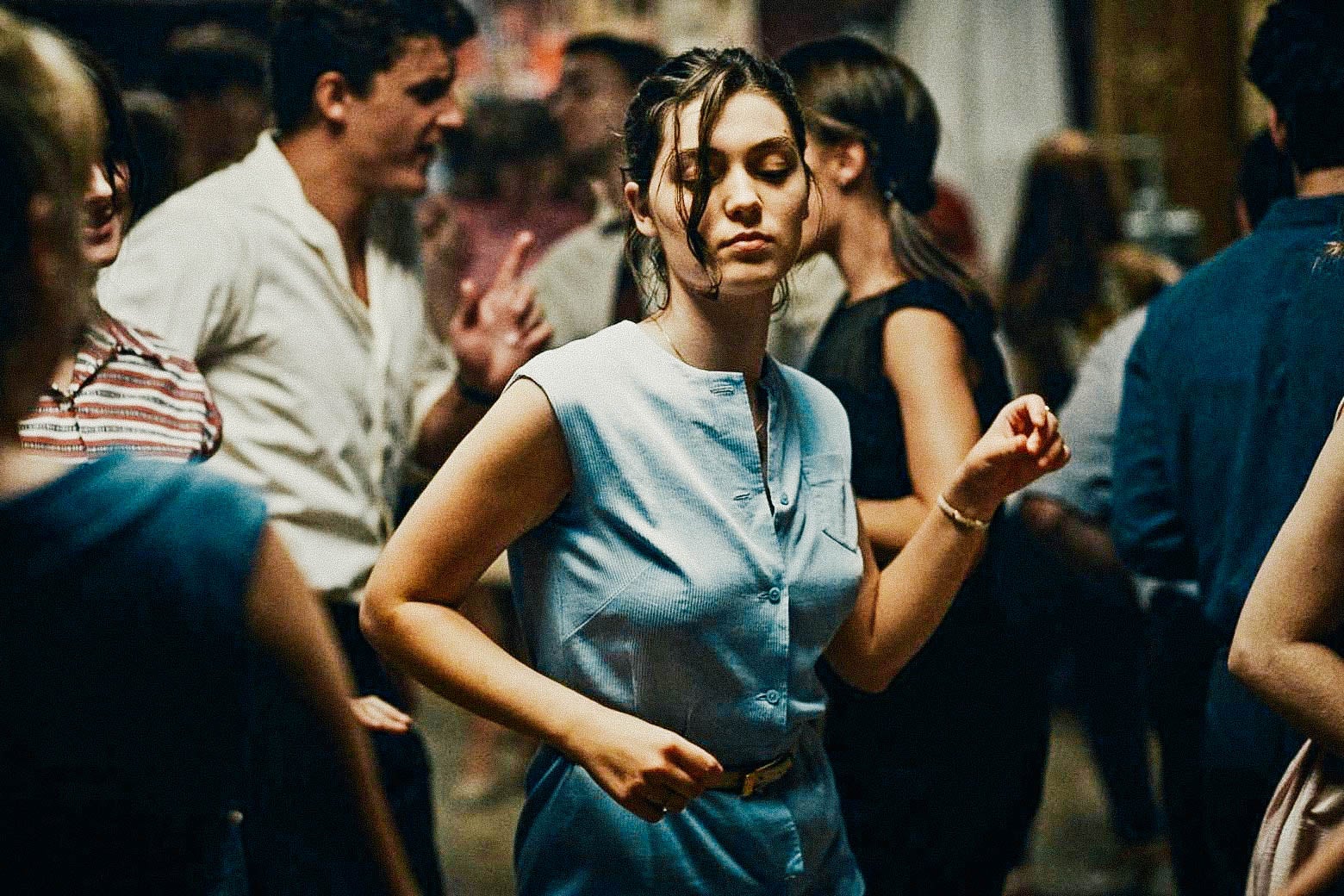 A slim young woman with fair skin and brown bangs and a blue 1960s dress dances on her own among a crowd of fellow young people, her eyes cast down.