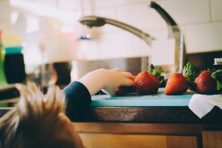A child reaches onto the counter to grab strawberries from a cutting board.