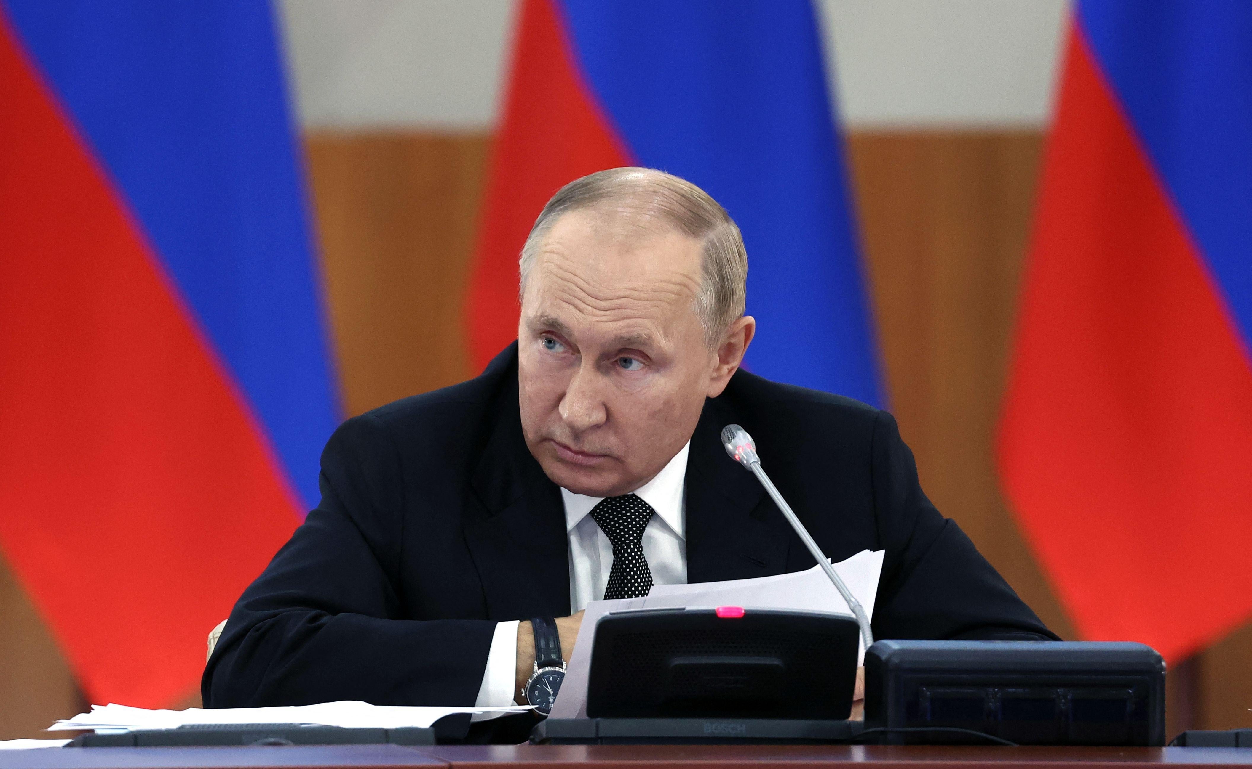 Russian President Vladimir Putin chairs a meeting of the State Council Presidium on the development of the national tourism industry at the Far Eastern Federal University in Vladivostok on September 6, 2022. He is hunched over with Russian flags behind him.