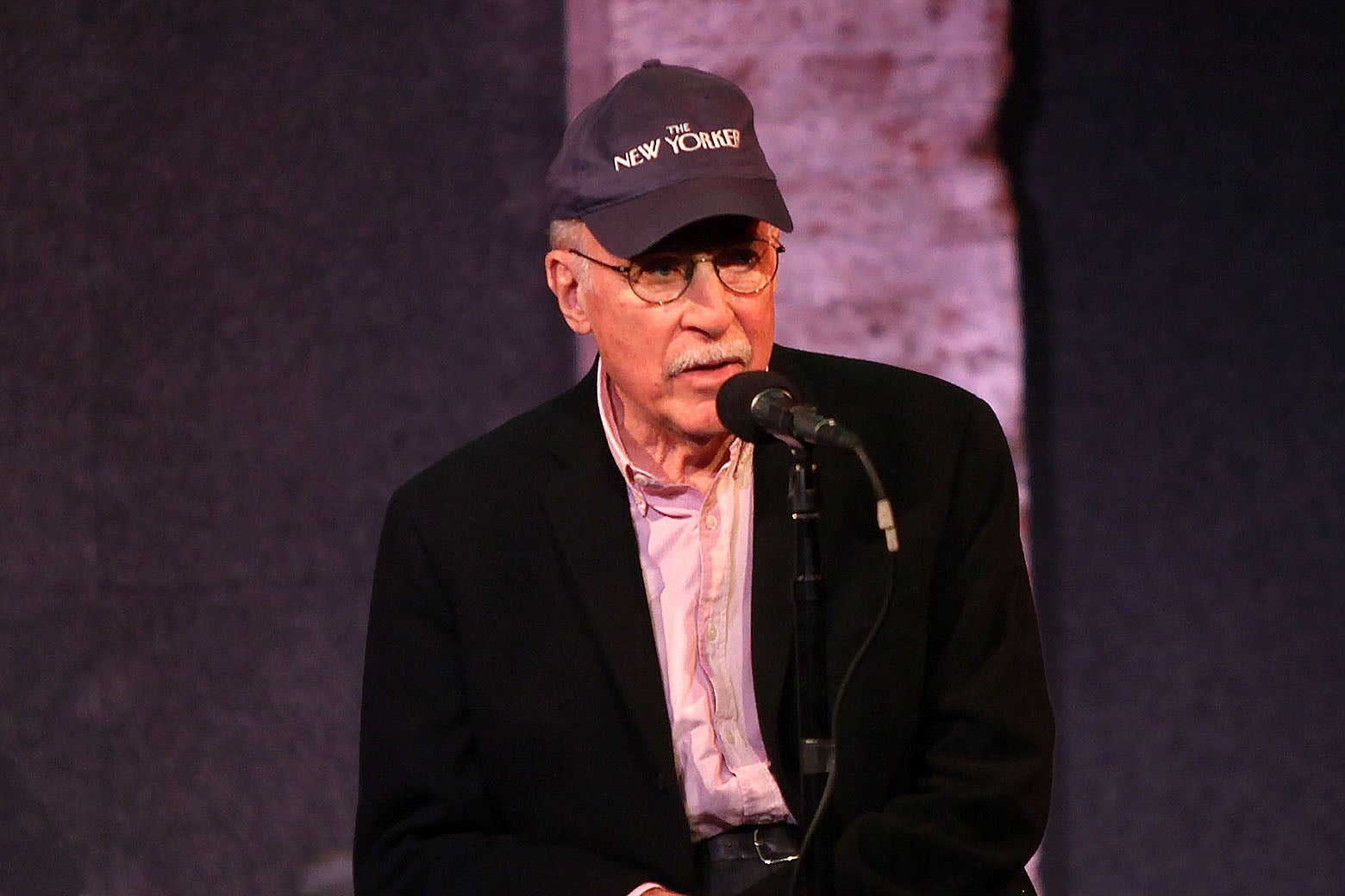 Roger Angell speaking to a crowd with a New Yorker cap on.