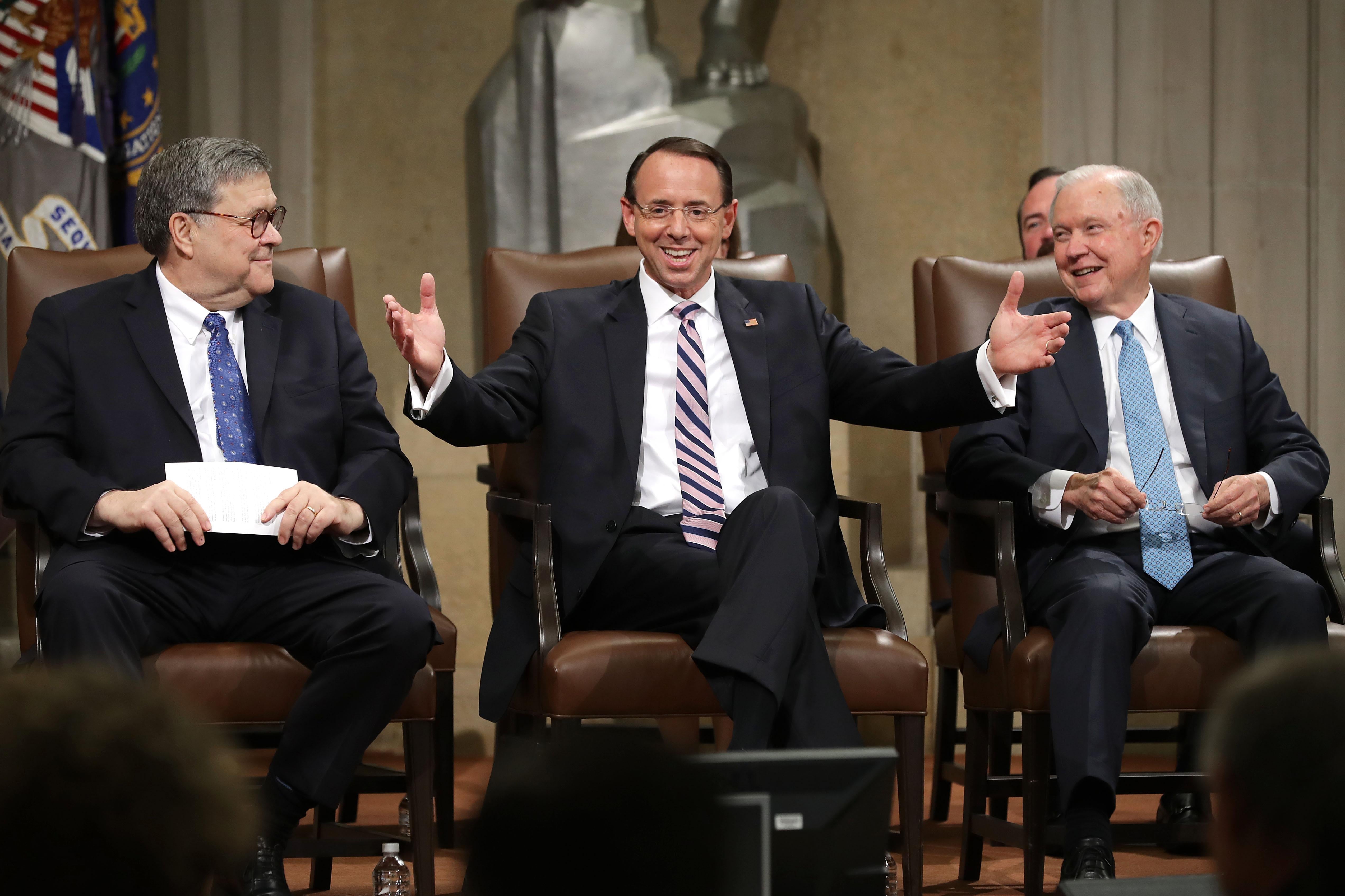 Rod Rosenstein smiles and raises his arms to a crowd as he sits onstage between Bill Barr and Jeff Sessions, both also smiling.