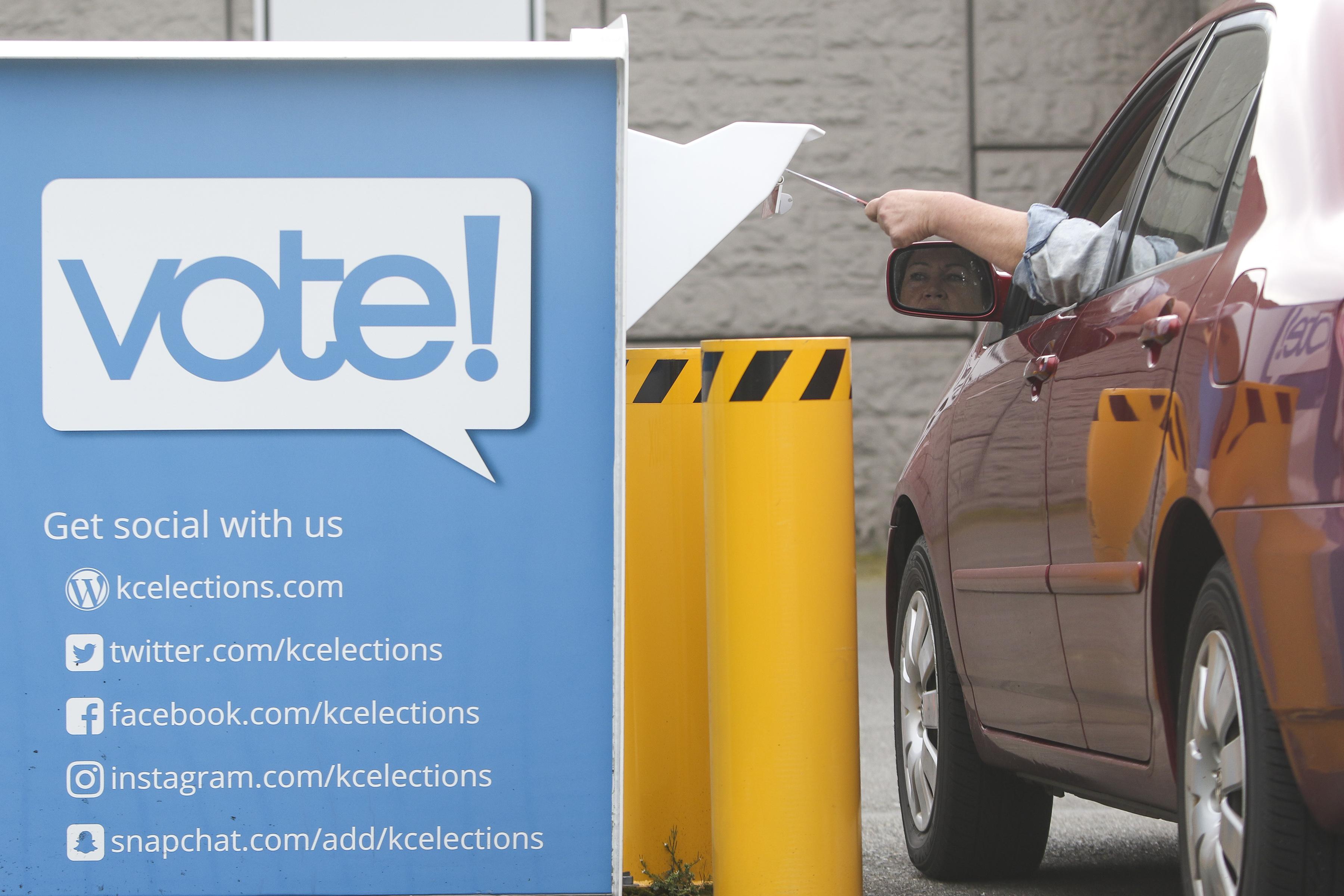 A person in a car puts a ballot in a drop box next to a sign listing local election social media accounts.