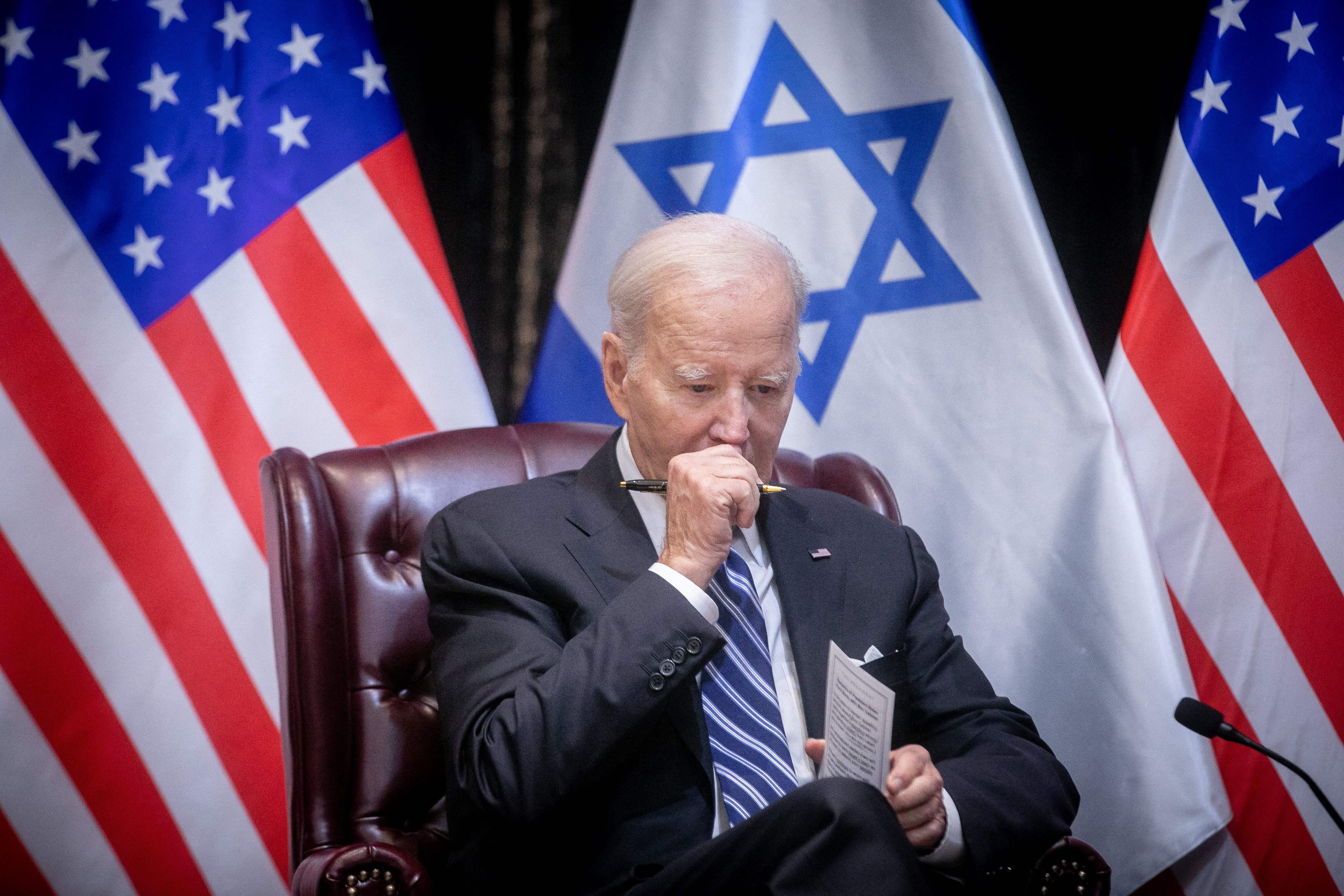Biden seated in front of Israeli and American flags, looking downward with his fist clutched to his mouth, holding a pen, and a notecard or pamphlet of some kind in his other hand.